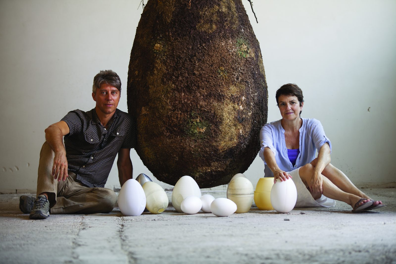 Two people seated beside a large earth-covered egg-shaped Capsule, with smaller pale egg-shaped forms arranged in front